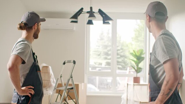 Two Caucasian male workers talking in bright apartment during renovation. Bearded man smiling, colleague adjusting cap. Technicians in overalls communicating indoors near window and tools.