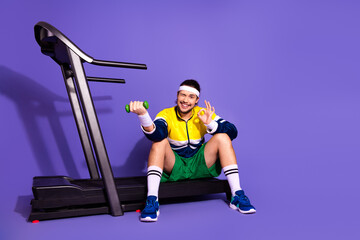 Young man in retro sportswear sitting on treadmill with dumbbell, making ok hand gesture against...