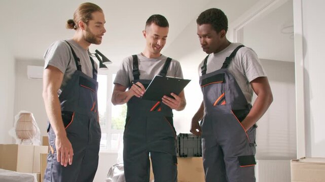Multiracial team of three male workers discussing checklist in modern apartment. One pointing at clipboard, others listening carefully. Men in overalls planning task during renovation indoors.