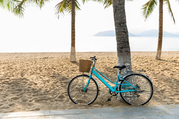 Fototapeta premium A retro-style blue bicycle with straw basket parked near palm trees on a sandy beach with blue sea or ocean background on the sunny morning. Tropical vacation concept.