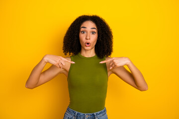 Portrait of a cheerful young woman with curly black hair on a vibrant yellow background pointing to...