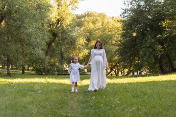 A pregnant woman and her daughter are dressed in white clothes and walking in the park. A happy...