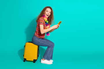 Cheerful young woman with curly hair sitting on luggage using a smartphone against turquoise...