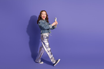 Cheerful schoolgirl with stylish denim outfit and metallic pants taking a smartphone photo on a...