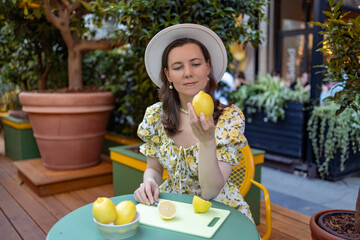 A woman in a floral dress and hat sits at a table outdoors, holding a lemon, with a bowl of lemons and a cutting board in front of her