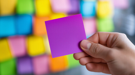 A hand holds a vibrant pink sticky note against a colorful background of many blurred sticky notes.
