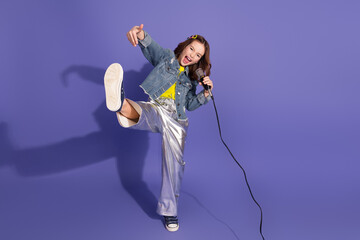 Joyful girl in casual trendy outfit singing with a microphone on a vibrant purple background...