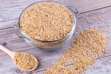 Glass bowl of uncooked brown rice with wooden spoon on rustic wooden table
