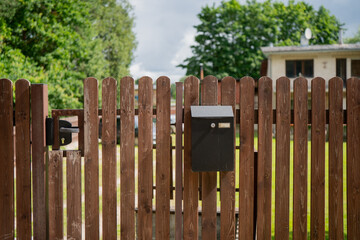Brown wooden picket fence with black mailbox and closed gate in front of residential property,...