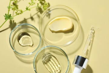 Different cosmetic products in Petri dishes, dropper and floral decor on pale yellow background,...