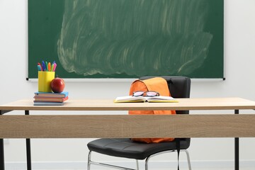 Different school stationery, glasses and red apple on wooden table near blackboard indoors