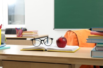 Different school stationery, glasses and red apple on wooden table near blackboard indoors