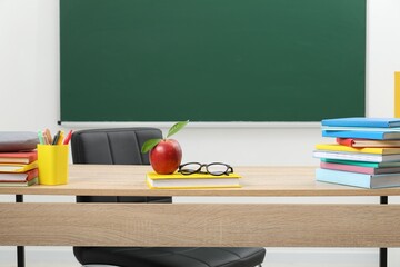 Different stationery, glasses and red apple on wooden table near blackboard indoors