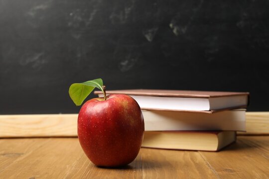Different books and red apple on wooden table near blackboard