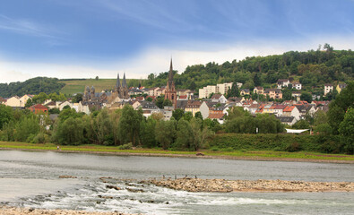 Pretty German Village viewed from a river cruise boat, on the bank of The Rhine River, with church steeple amongst pretty stone houses nd lush forest backdrop.