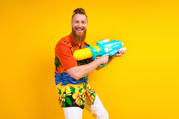 Young man with tropical shirt holding water gun posing against yellow background celebrating summer...