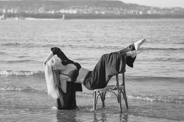 Girl in a black business suit in the sea on the beach on a wooden retro chair. Photo model....