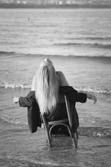 Girl in a black business suit in the sea on the beach on a wooden retro chair. Photo model....