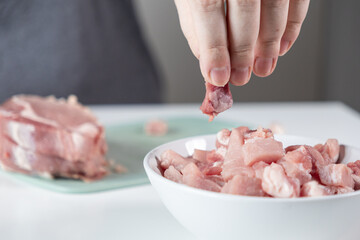 A person's hand carefully places fresh raw pork meat pieces into a white bowl, illustrating the...