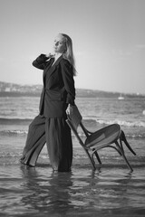 Girl in a black business suit in the sea on the beach on a wooden retro chair. Photo model. Fashion. Posing. Black and white photo