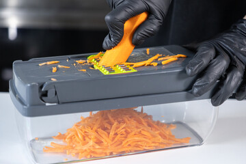 Person in black protective gloves expertly grates a fresh orange carrot using a modern mandoline...