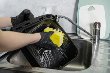 A person wearing protective black gloves diligently cleans a greasy oven baking tray with a soapy yellow sponge, promoting excellent kitchen hygiene and household cleanliness