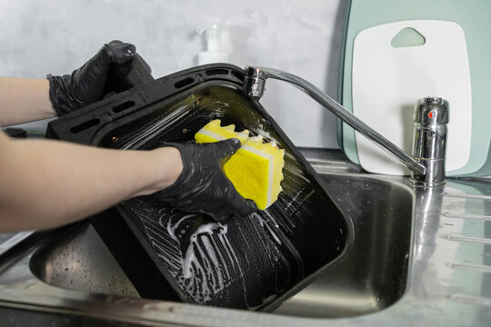 A person wearing protective black gloves diligently cleans a greasy oven baking tray with a soapy yellow sponge, promoting excellent kitchen hygiene and household cleanliness