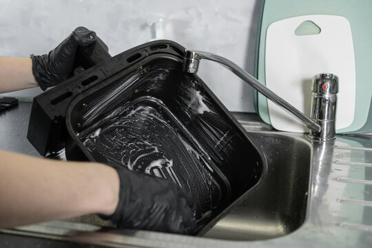 Person in black gloves washing a dirty air fryer basket with soap suds in a modern kitchen sink, demonstrating home cleaning and appliance upkeep