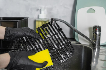 Hands in black gloves diligently cleaning an air fryer grill grate with a yellow sponge and soap in...