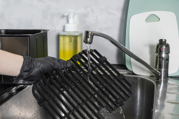 Hand wearing a black protective glove washes a dirty non-stick air fryer grill grate under running...