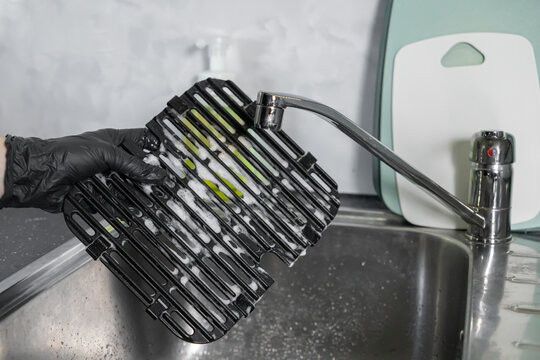Person wearing a black glove diligently washing a dirty air fryer grill grate covered in foamy soap suds within a clean stainless steel kitchen sink, showcasing essential domestic hygiene