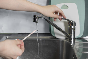 Person's hand holding a water testing strip under running kitchen faucet, analyzing home tap water for drinking safety and quality control