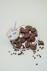 Iced coffee drink with a colorful straw beside a silver plate filled with chocolate cookies shaped like coffee beans, surrounded by scattered coffee beans on a light background