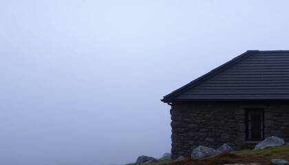 A single line of warm light outlines the corner of a stone-clad cabin, barely piercing the quiet veil of coastal mist.
