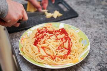 On the table in the kitchen there is a plate of spaghetti with ketchup on it near the plate a woman is cutting cheese on a cutting board. With a knife.
