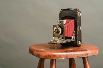 An old antique leather accordion camera, about 100 years old, stands on a wooden chair the background is gray paper.