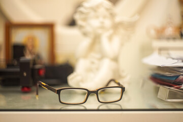 A pair of square reading glasses lies on a table covered with glass, and there are papers to the right and in the corner and Household items are visible in the blurred background