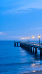 Obraz premium A long pier with glowing lampposts extends into a calm, blue sea under a twilight sky.