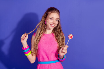 Charming young woman in pink dress enjoying leisure time with a lollipop against a vibrant purple...