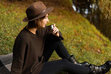 A woman enjoys a warm cup of tea during a serene autumn picnic by a peaceful lake, surrounded by fallen leaves and golden light.