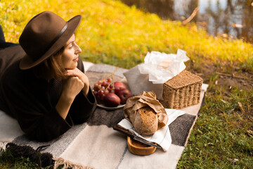 Golden leaves surround a woman in a hat resting on a blanket at a fall picnic, enjoying fruit and...