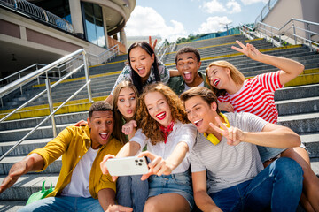 Group of cheerful young friends taking a selfie outdoors on urban steps during a sunny day,...