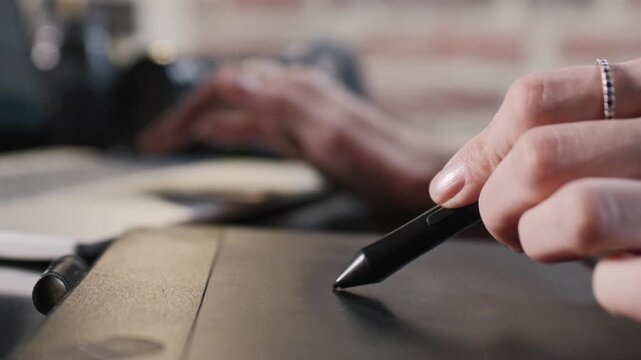 Caucasian hand holding stylus drawing on graphic tablet near laptop, creating digital art at home, focus on pen touching surface while other hand working in background on keyboard. Close up.