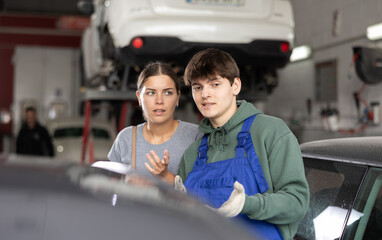 Interested female client talking to young auto mechanic, discussing needed maintenance of her car...