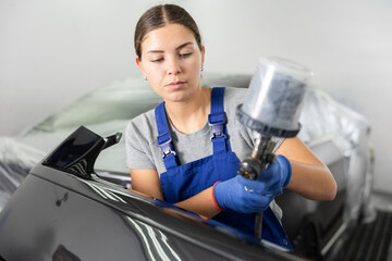 Young woman mechanic in uniform paints car with machine in car service station
