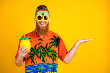 Bearded man in tropical shirt gestures cheerfully while holding pineapple-shaped cup against yellow...