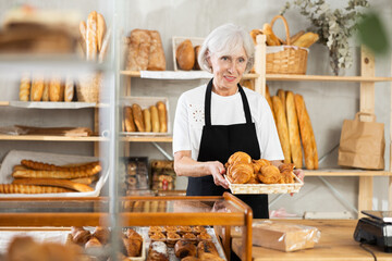 Friendly female baker in apron putting hot delicious croissants to the showcase for sale