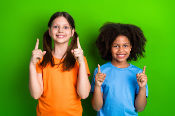 Two cheerful girls in colorful outfits pointing upward with cheerful smiles against green background