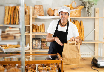 Positive middle-aged salesman giving paper bag containing baked goods in bakery shop