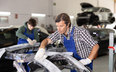 Experienced auto body technician in blue overalls focused on smoothing surface of car parts using...